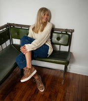 A woman with long blonde hair sits on a green bench, wearing a cream sweater, blue jeans, and the Unwavering sneakers by Bed|Stü, smiling and looking to the side.