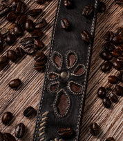 Upswing II dark leather belt strap with a stitched flower cutout and center rivet, displayed as an artisanal floral design amidst scattered coffee beans on a wooden surface.