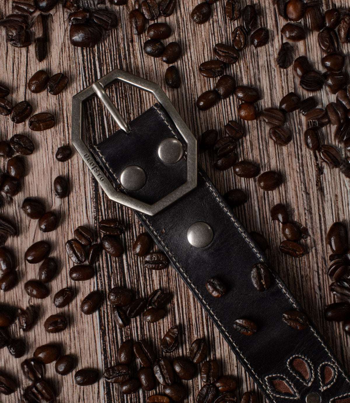 Bed|Stü Upswing II dark, handcrafted leather belt with a hexagonal silver buckle, resting on a surface scattered with roasted coffee beans.