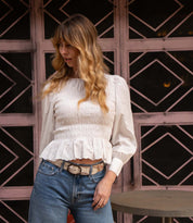 A woman with long wavy hair wears a white smocked blouse, blue jeans, and the Bed Stu Upswing II artisanal belt as she stands outdoors near a round table and a geometric window.