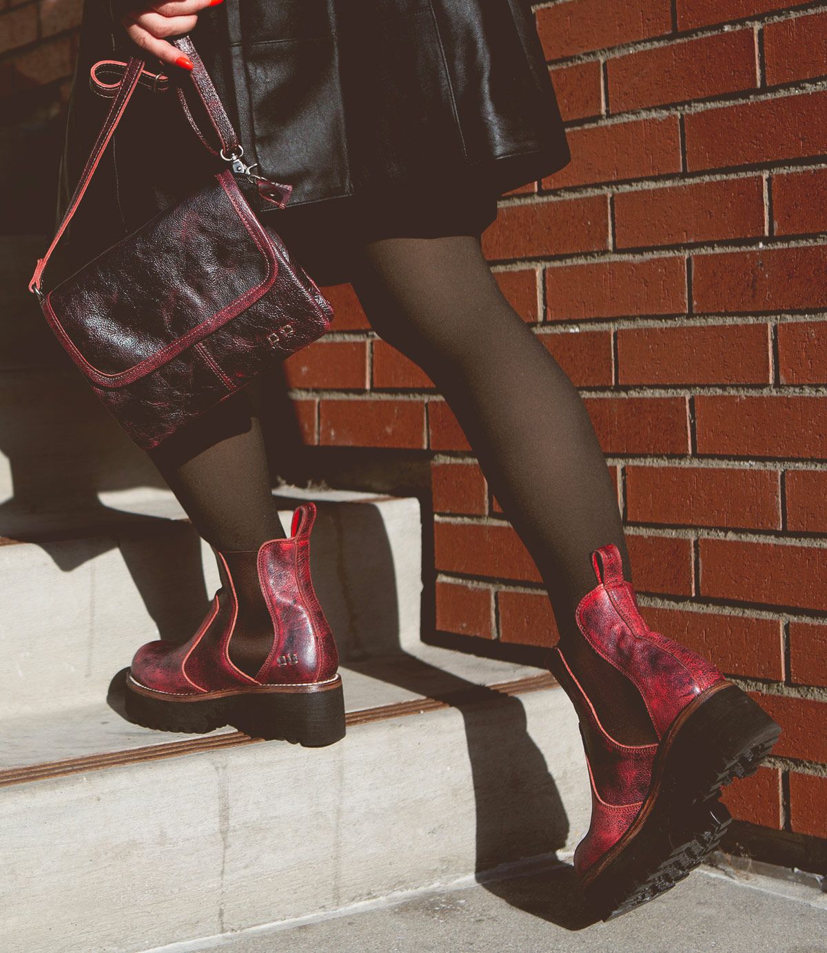 Person wearing black tights, red Valda Hi Chelsea boots from Bed Stu with a platform sole, and a black leather skirt, holding a black handbag while walking up concrete stairs next to a brick wall.