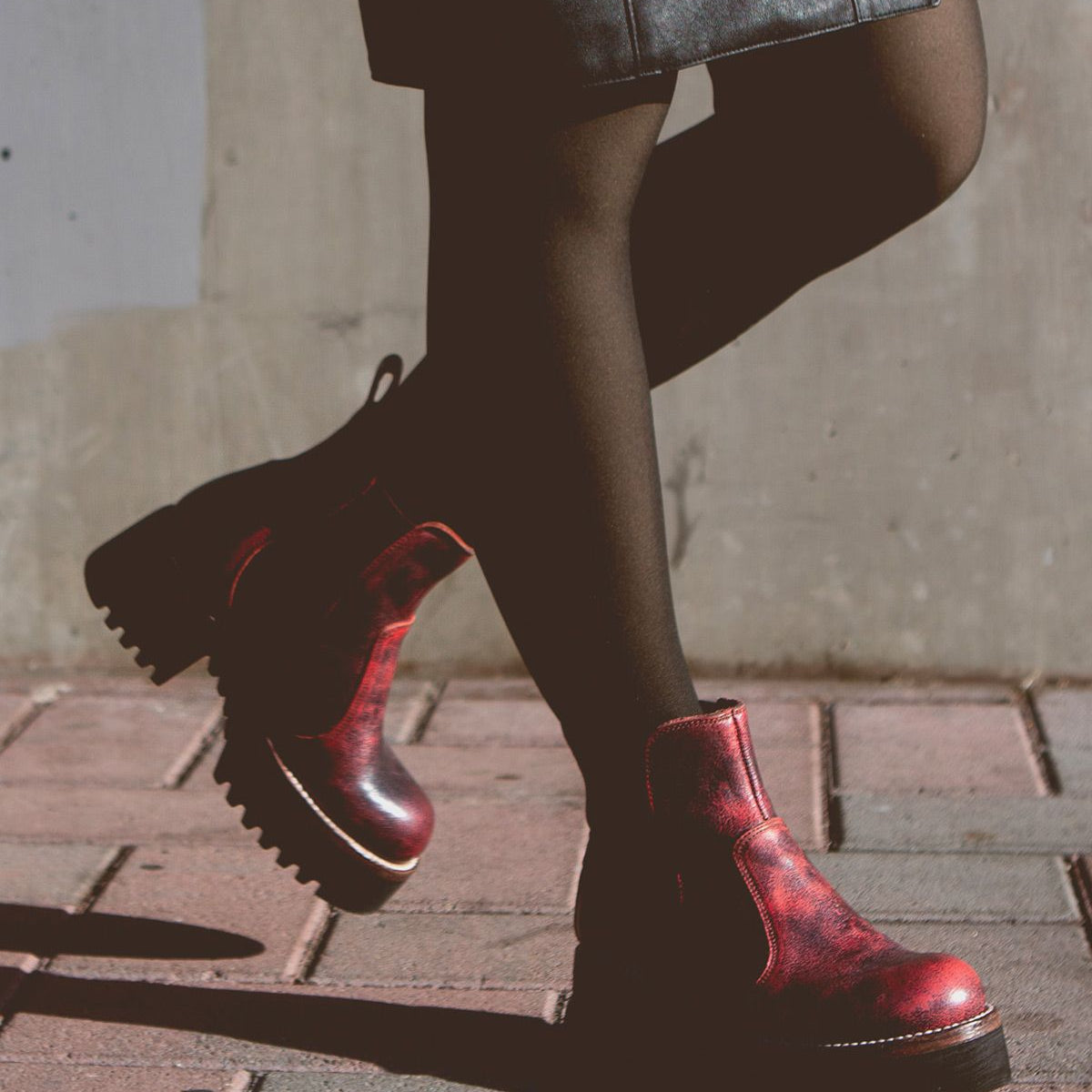 Person wearing black tights, a leather skirt, and Bed Stu's Valda Hi red ankle boots with a platform sole walking on a brick pathway against a concrete wall.