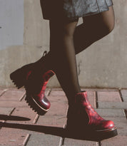 Person wearing black tights, a leather skirt, and Bed Stu's Valda Hi red ankle boots with a platform sole walking on a brick pathway against a concrete wall.