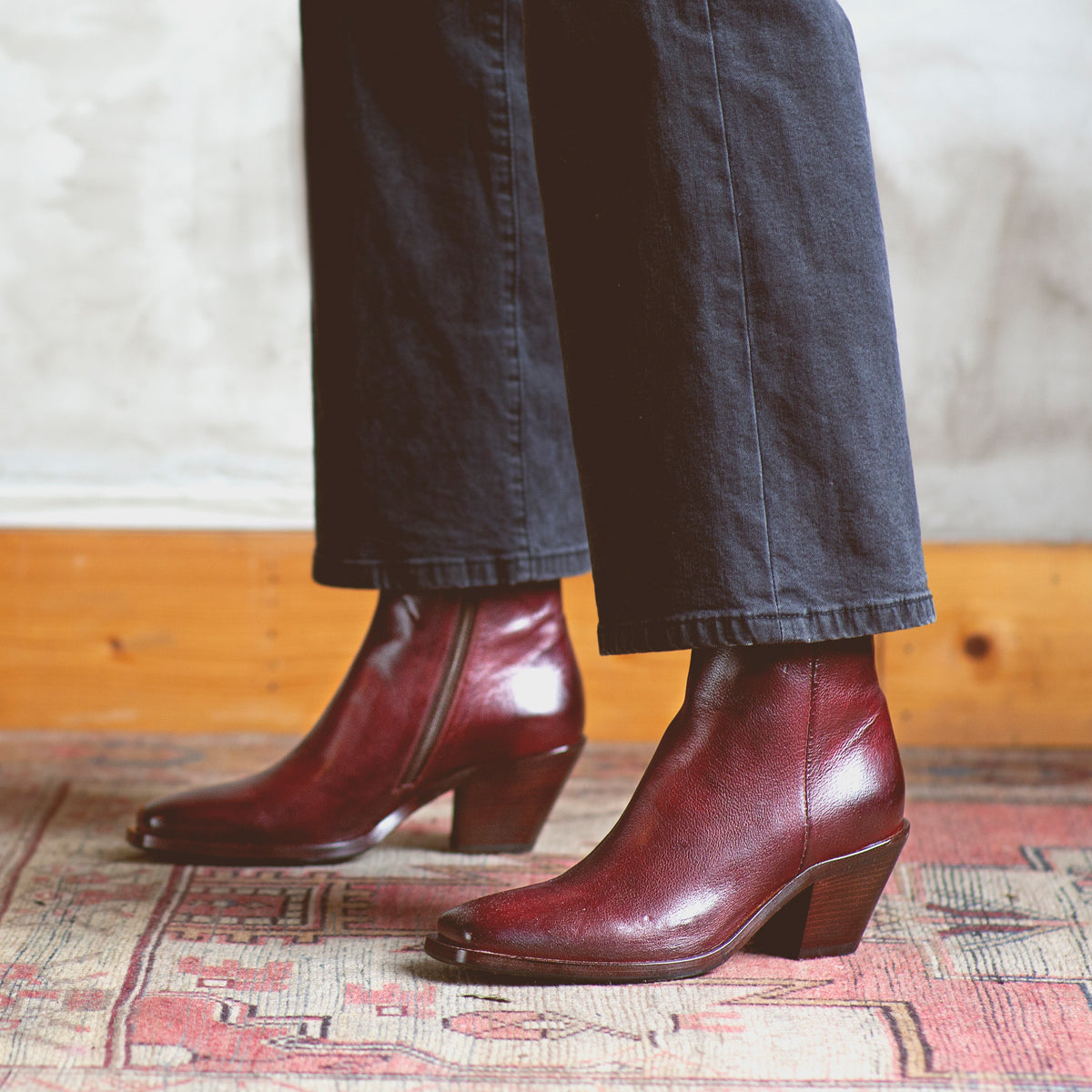 A woman showcasing a pair of Bed Stu Vendue burgundy boots with a sleek silhouette and square toe on a rug.