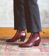 A woman showcasing a pair of Bed Stu Vendue burgundy boots with a sleek silhouette and square toe on a rug.