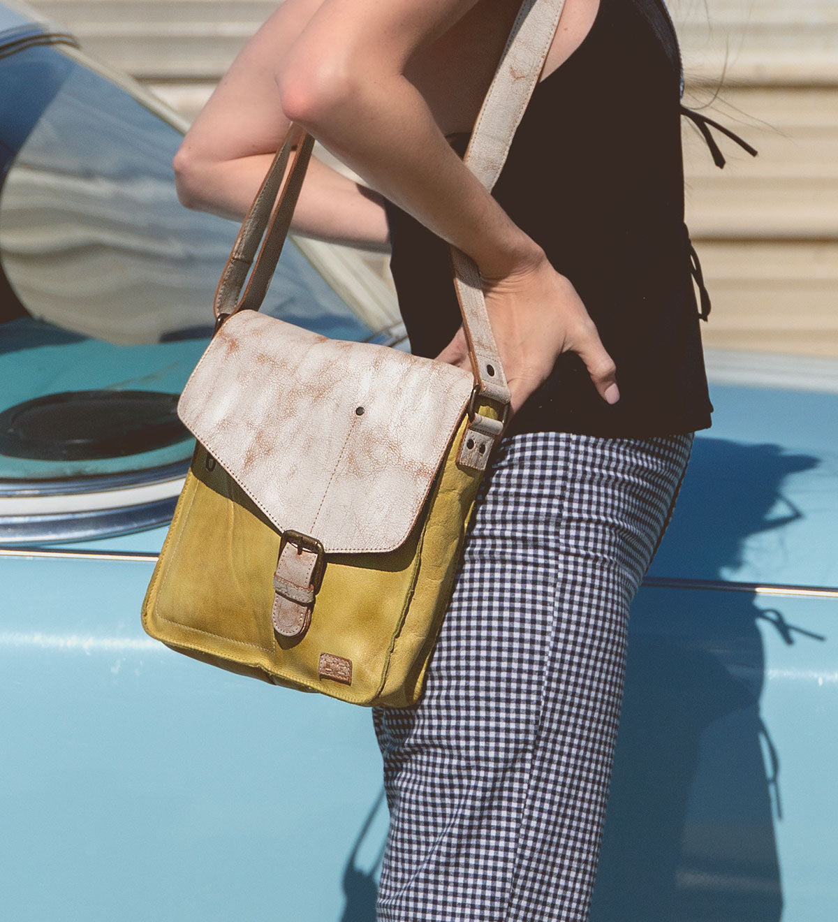A person leans against a blue car, wearing a black top and checkered pants, holding the Venice Beach handmade leather crossbody bag by Bed Stu, featuring a bohemian-inspired design.