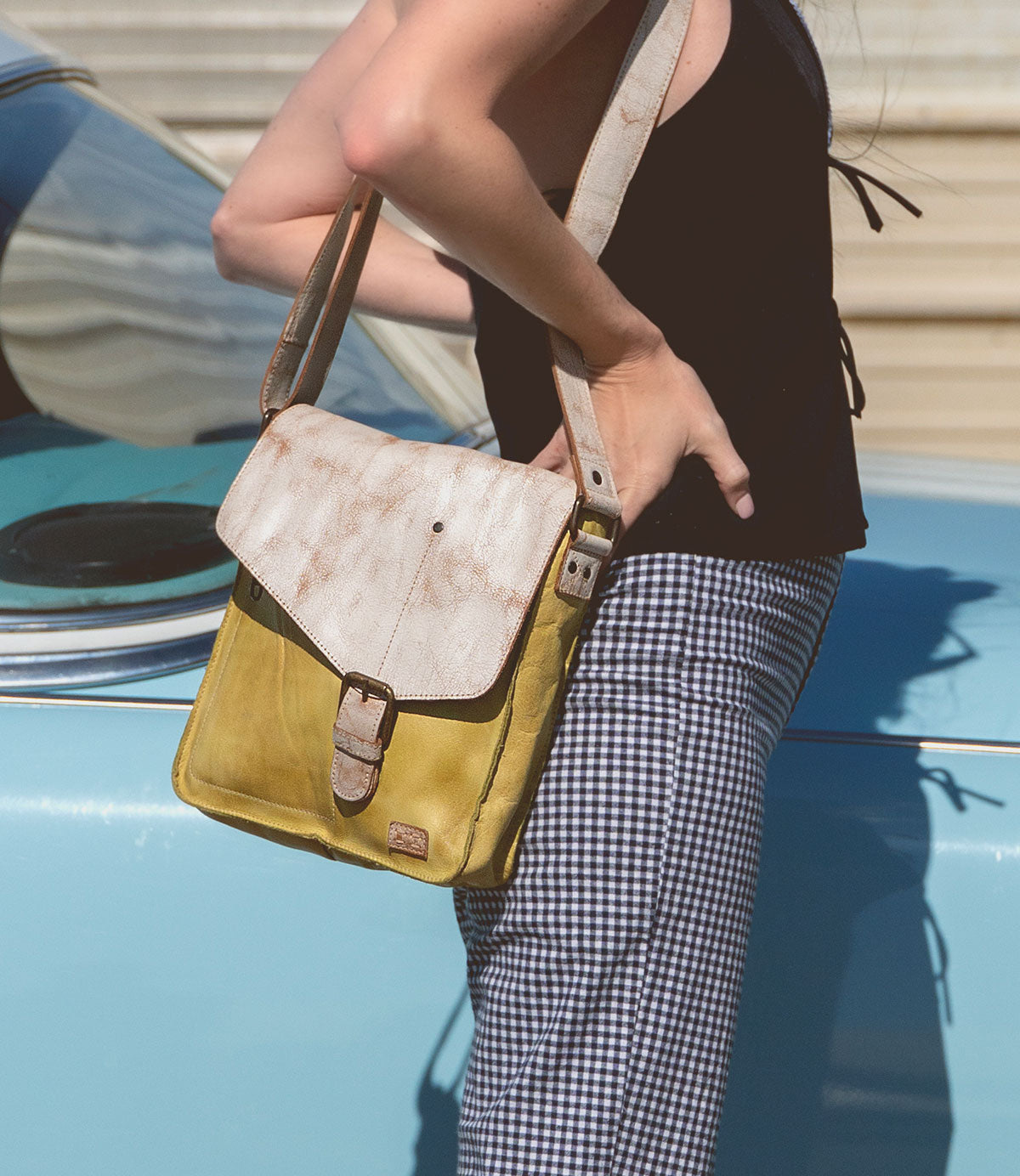 A person leans against a blue car, wearing a black top and checkered pants, holding the Venice Beach handmade leather crossbody bag by Bed Stu, featuring a bohemian-inspired design.