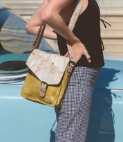 A person leans against a blue car, wearing a black top and checkered pants, holding the Venice Beach handmade leather crossbody bag by Bed Stu, featuring a bohemian-inspired design.
