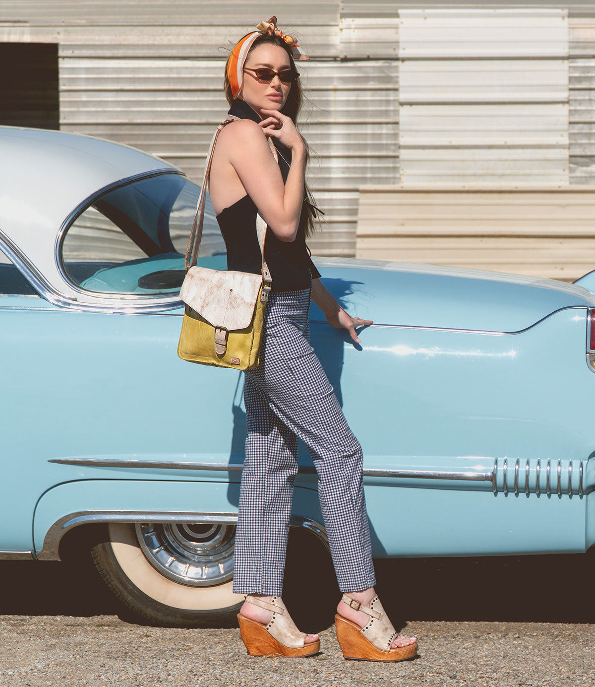 A woman in sunglasses and a headscarf stands by a vintage blue car, wearing a black top, checkered pants, wedge sandals, and carrying Bed Stu's Venice Beach handmade leather crossbody bag that showcases quality craftsmanship.