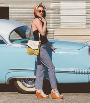 A woman in sunglasses and a headscarf stands by a vintage blue car, wearing a black top, checkered pants, wedge sandals, and carrying Bed Stu's Venice Beach handmade leather crossbody bag that showcases quality craftsmanship.