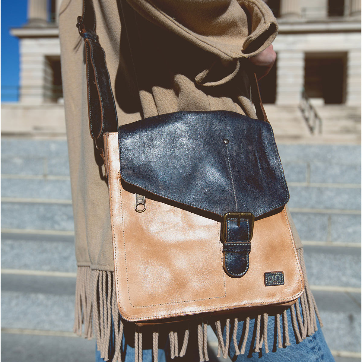 A person wearing a beige coat with fringes carries the Venice Beach, a bohemian and vintage-inspired masterpiece by Bed Stu—a tan and black handmade leather crossbody bag—while walking on city steps.