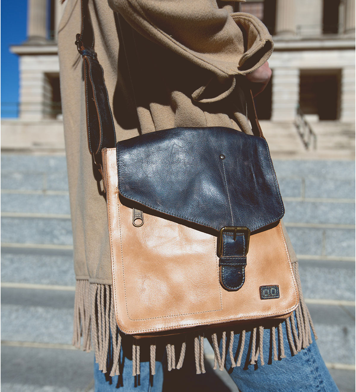 A person wearing a beige coat with fringes carries the Venice Beach, a bohemian and vintage-inspired masterpiece by Bed Stu—a tan and black handmade leather crossbody bag—while walking on city steps.