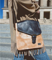 A person wearing a beige coat with fringes carries the Venice Beach, a bohemian and vintage-inspired masterpiece by Bed Stu—a tan and black handmade leather crossbody bag—while walking on city steps.
