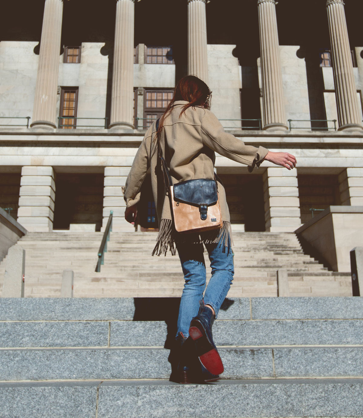 A person walks up stone steps towards a large building with columns, wearing a beige coat and jeans, while carrying the Venice Beach crossbody bag by Bed Stu, which exudes quality craftsmanship.