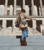 A person walks up stone steps towards a large building with columns, wearing a beige coat and jeans, while carrying the Venice Beach crossbody bag by Bed Stu, which exudes quality craftsmanship.