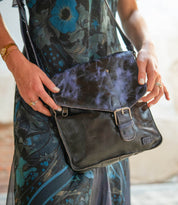 A person in a floral dress holds the Bed Stu Venice Beach handmade leather crossbody bag in black and purple, featuring a front buckle.