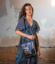 A woman stands against a worn, textured wall with peeling paint, carrying the Bed Stu Venice Beach handmade leather crossbody bag and wearing a blue patterned dress.