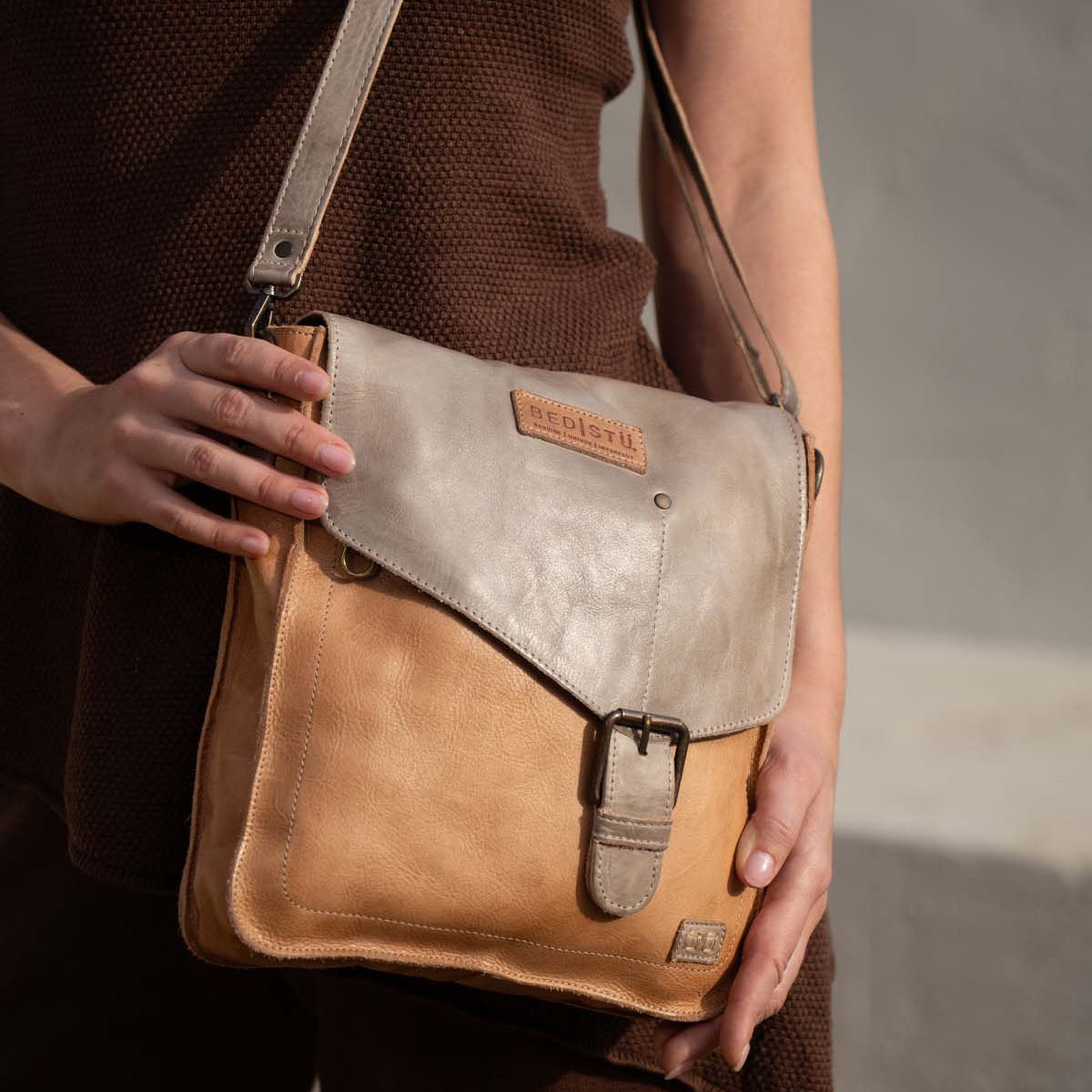 A person in a brown outfit holds the Venice Beach handbag by Bed Stu—a vintage-inspired, two-tone leather shoulder bag with a buckle and a visible brand patch.