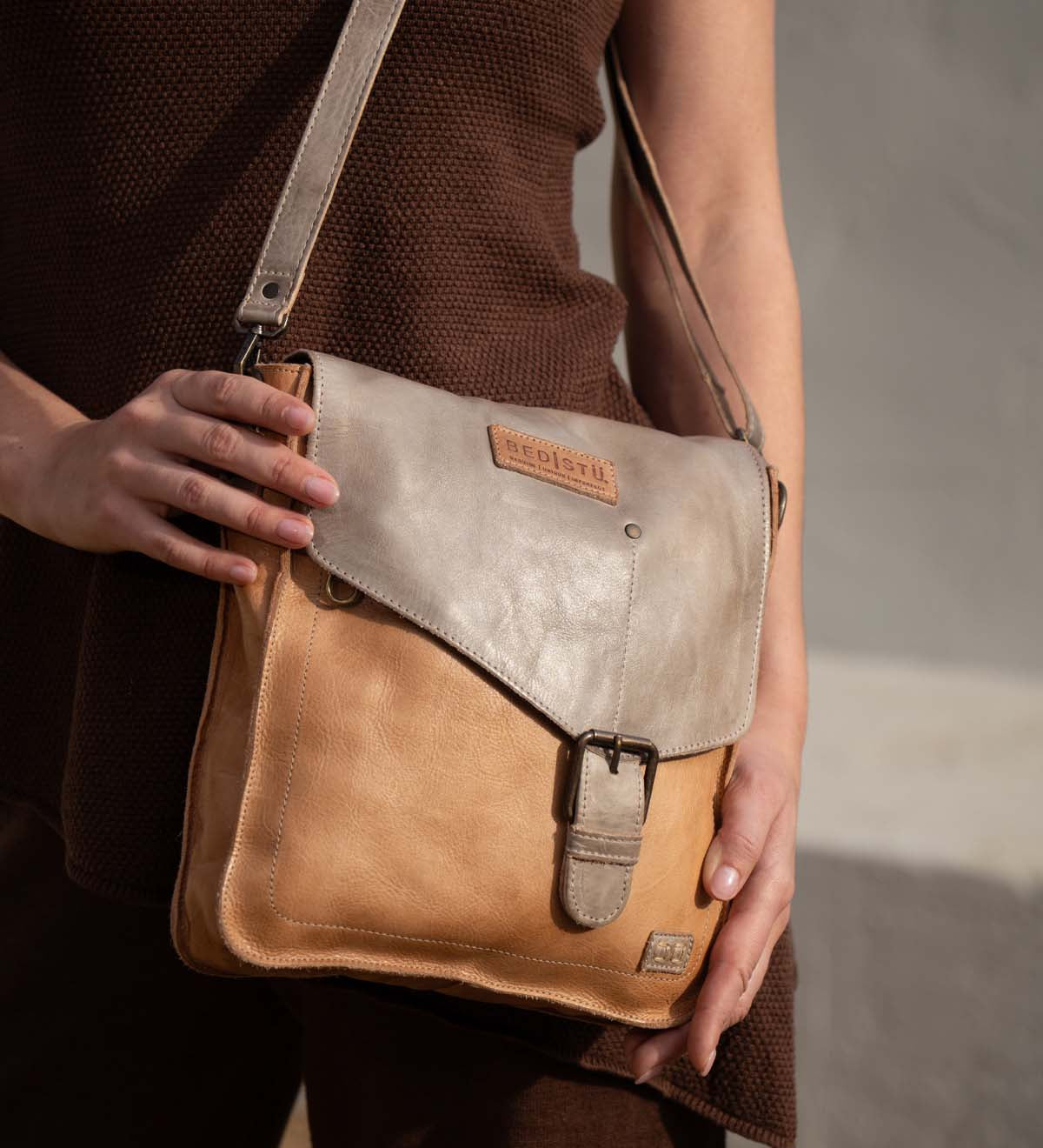 A person in a brown outfit holds the Venice Beach handbag by Bed Stu—a vintage-inspired, two-tone leather shoulder bag with a buckle and a visible brand patch.