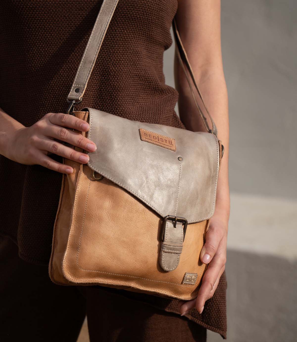 A person in a brown outfit holds the Venice Beach handbag by Bed Stu—a vintage-inspired, two-tone leather shoulder bag with a buckle and a visible brand patch.