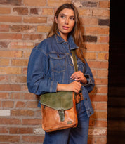 A woman in a denim jacket and jeans stands against a brick wall, holding the Bed Stu Venice Beach vintage-inspired brown and green leather handbag.