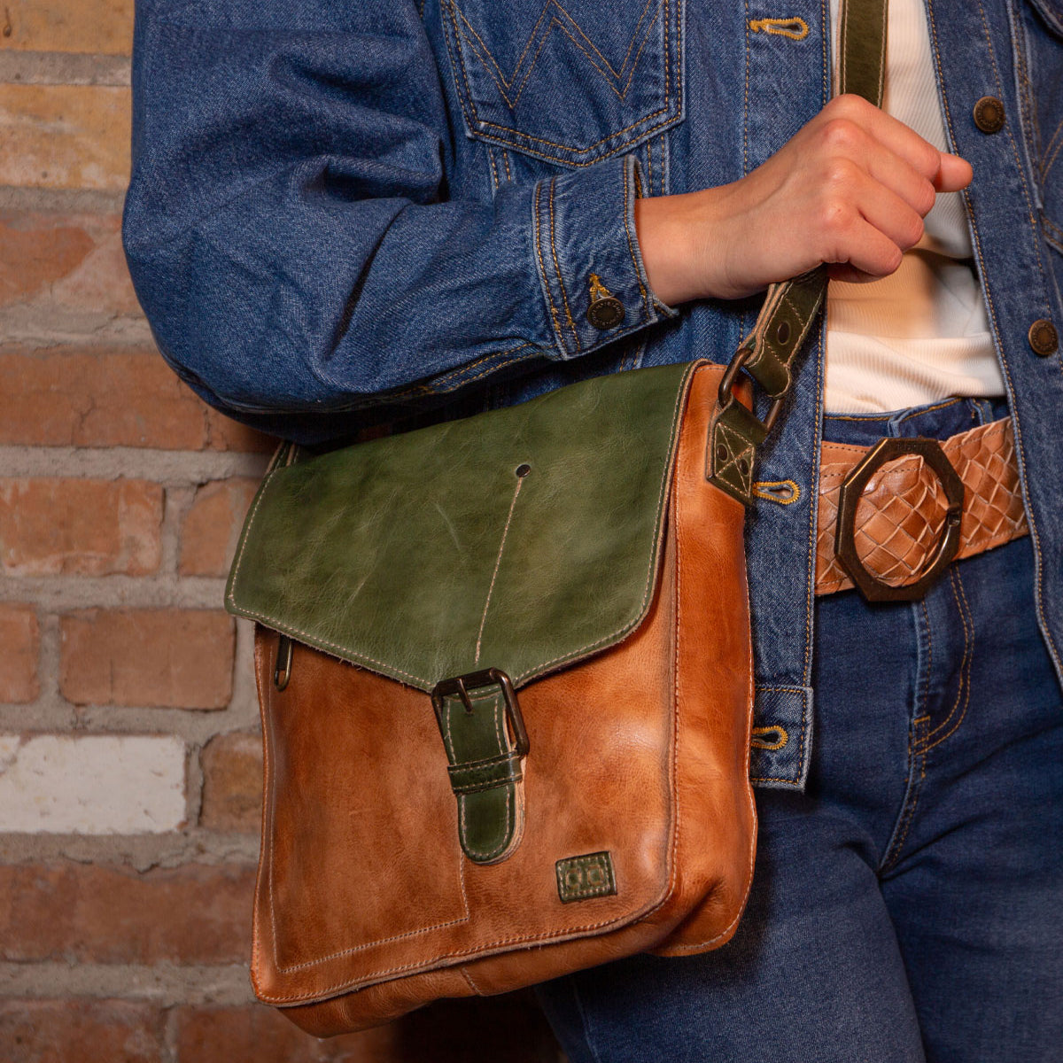 A person in a blue denim jacket and jeans with a tan belt holds the Bed Stu Venice Beach handmade leather crossbody bag in brown and green, standing before a brick wall.