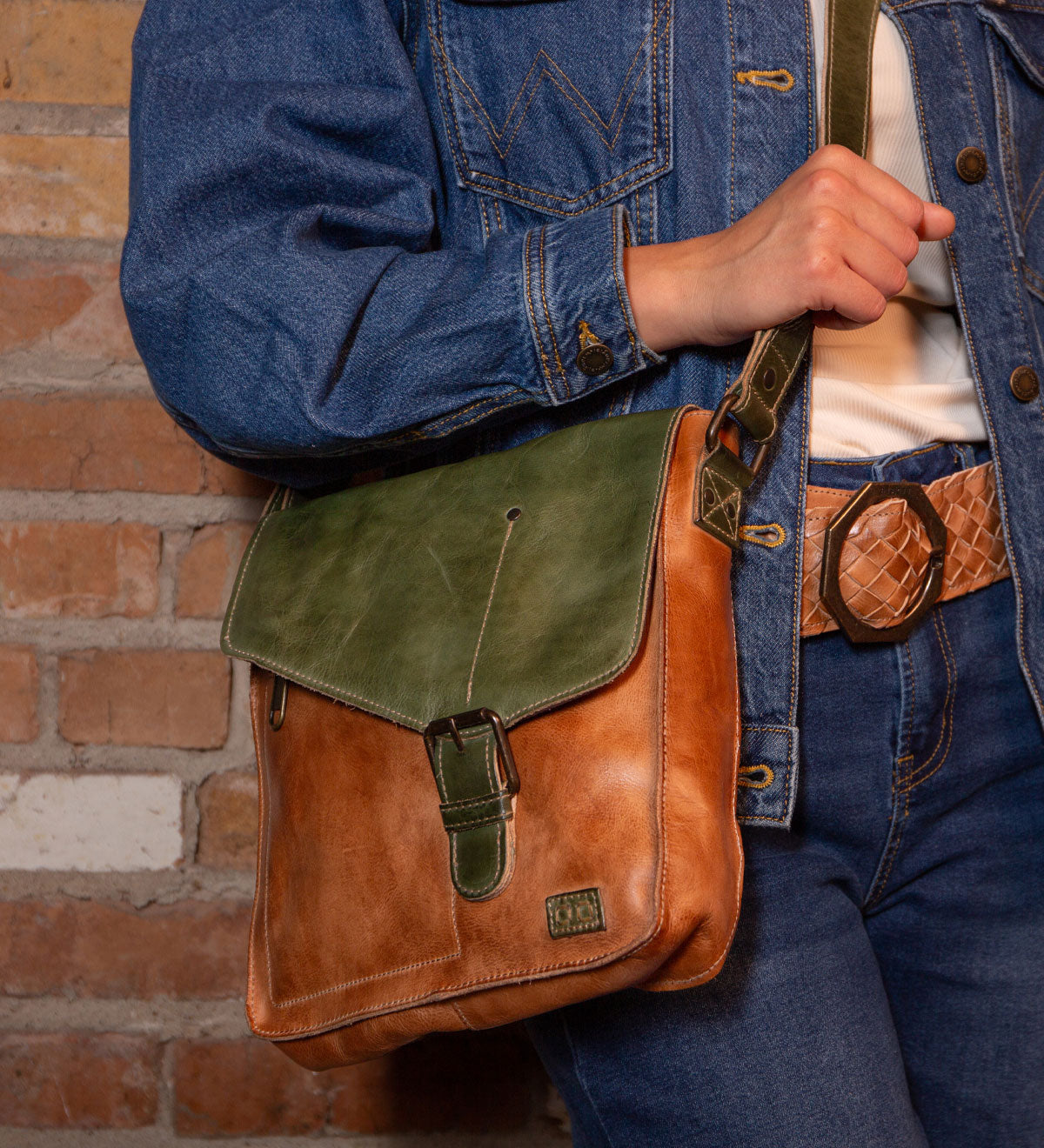 A person in a blue denim jacket and jeans with a tan belt holds the Bed Stu Venice Beach handmade leather crossbody bag in brown and green, standing before a brick wall.