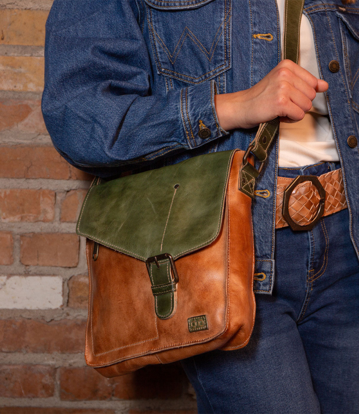 A person in a blue denim jacket and jeans with a tan belt holds the Bed Stu Venice Beach handmade leather crossbody bag in brown and green, standing before a brick wall.