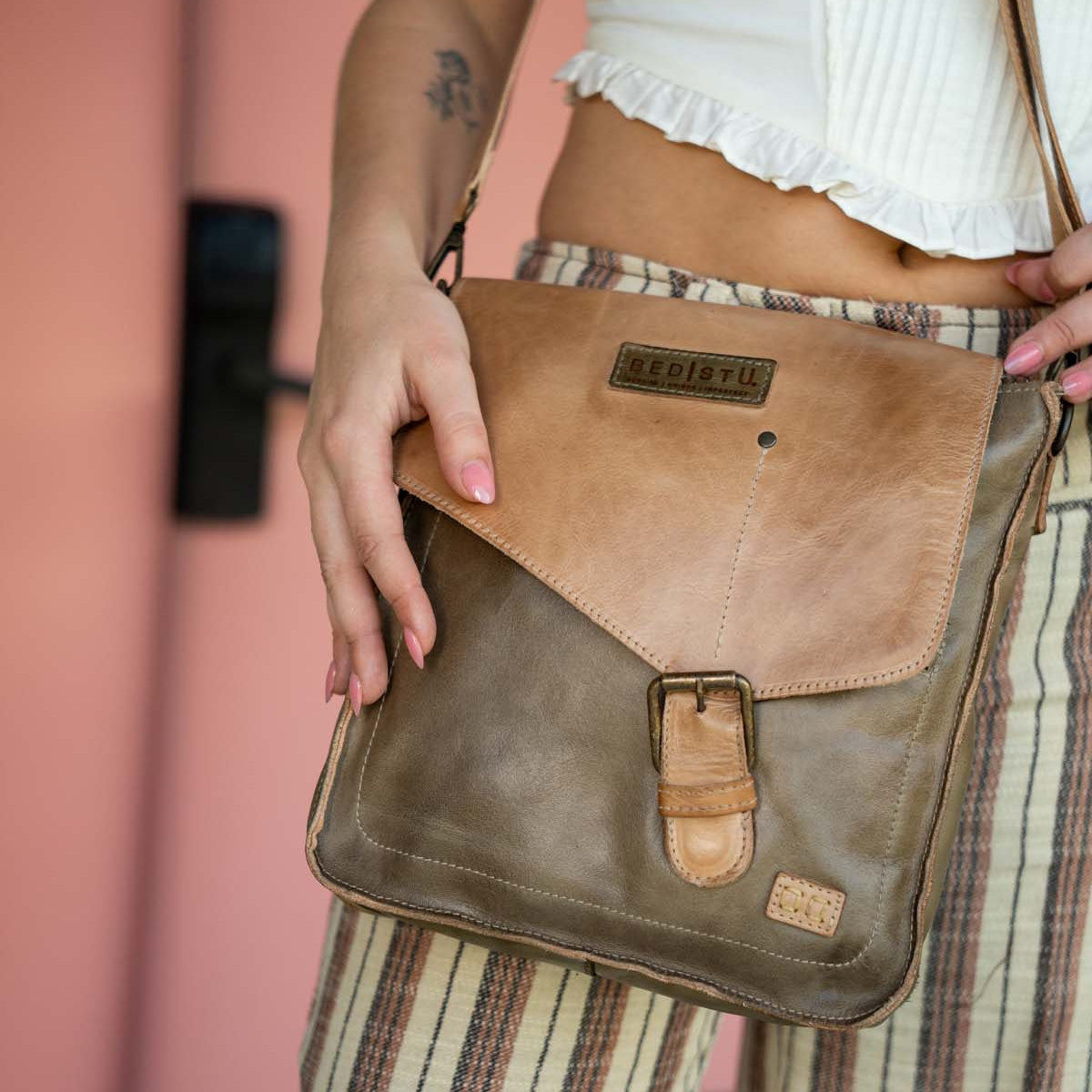Person wearing striped pants and a white top, holding the Bed Stu Venice Beach crossbody bag made from brown full-grain leather and featuring an adjustable strap, buckle, and branded label.