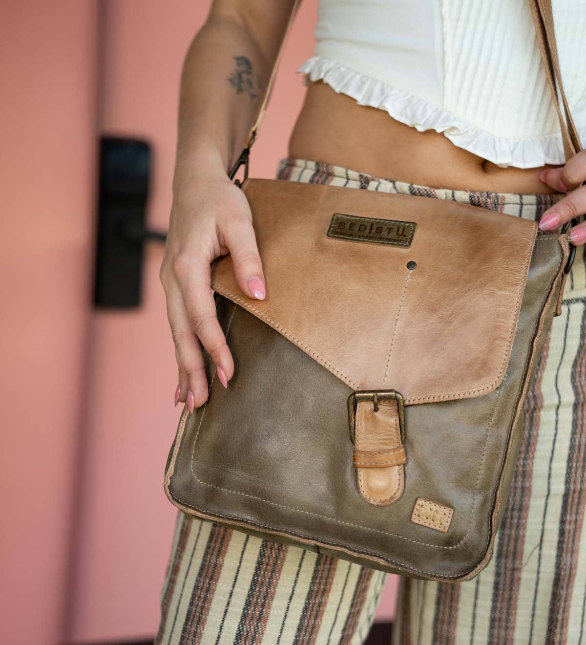 Person wearing striped pants and a white top, holding the Bed Stu Venice Beach crossbody bag made from brown full-grain leather and featuring an adjustable strap, buckle, and branded label.