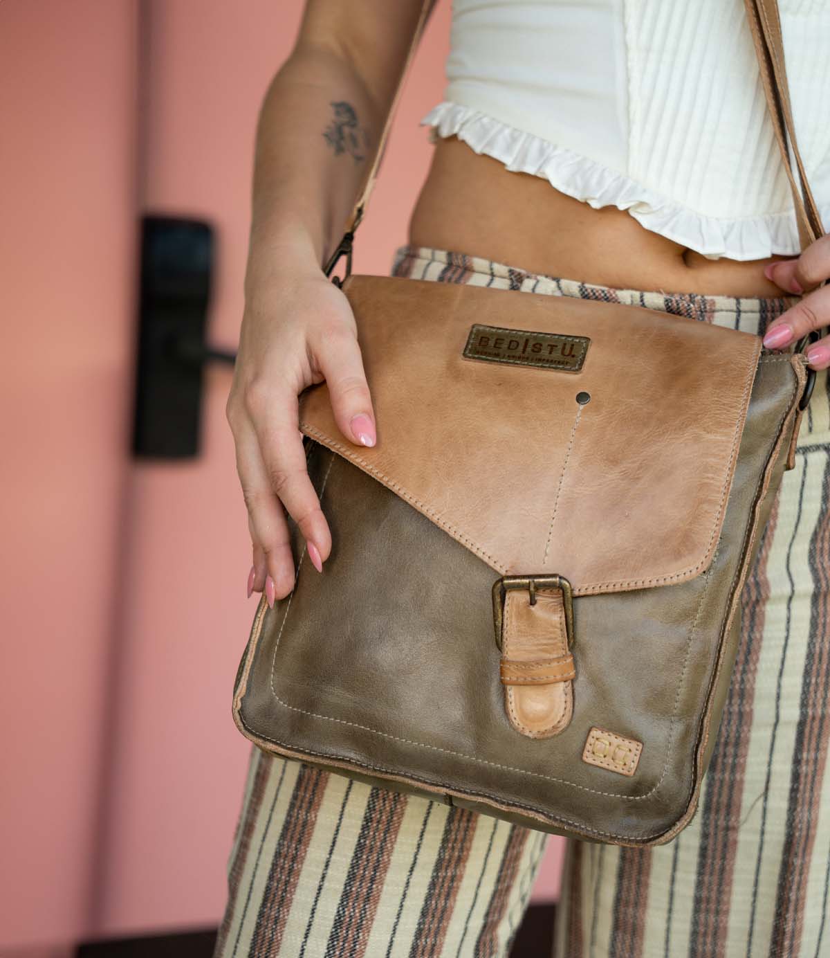 Person wearing striped pants and a white top, holding the Bed Stu Venice Beach crossbody bag made from brown full-grain leather and featuring an adjustable strap, buckle, and branded label.