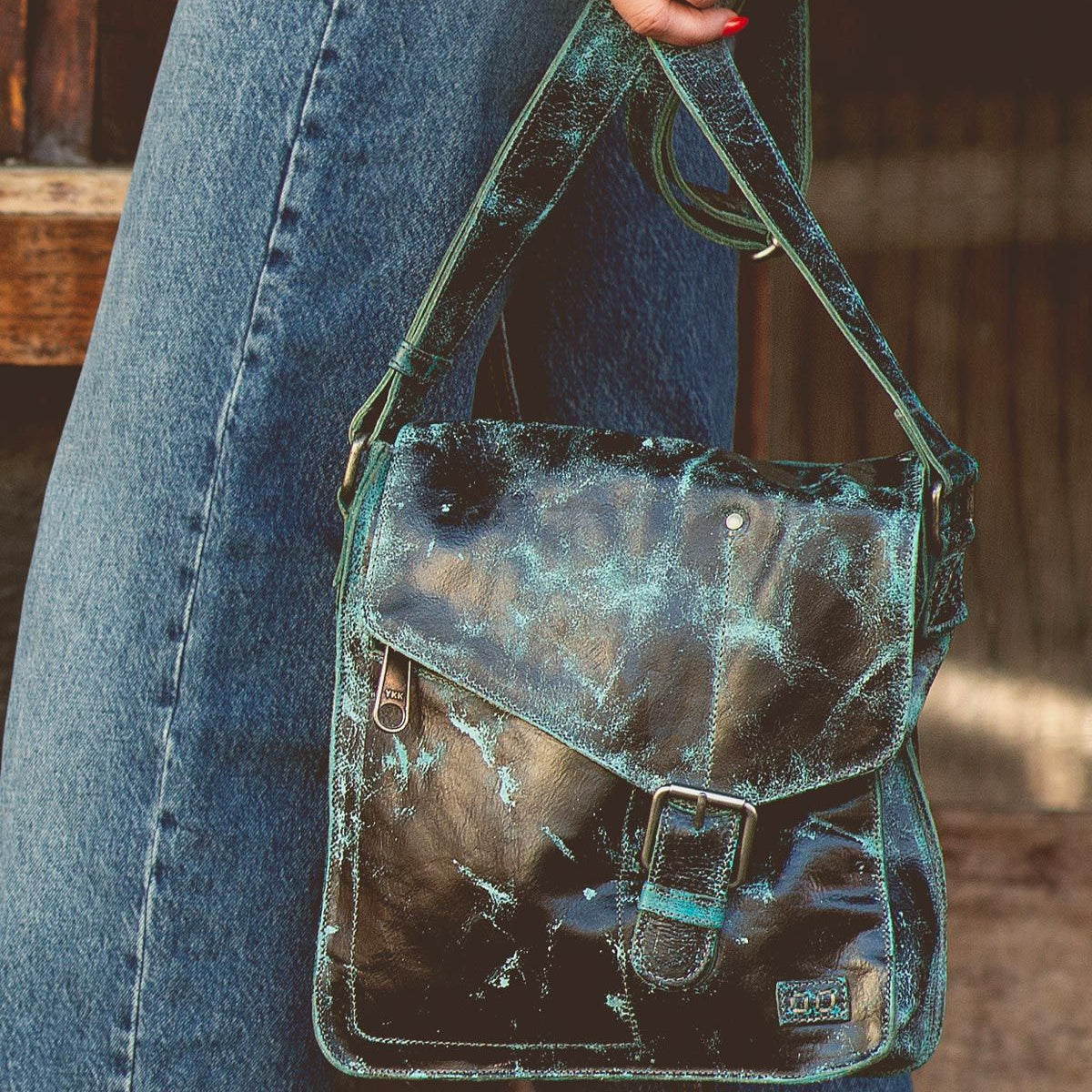 A person wearing blue jeans holds a Bed Stu Venice Beach handmade leather crossbody bag with an aged, distressed green and black leather finish and a buckle on the front flap.