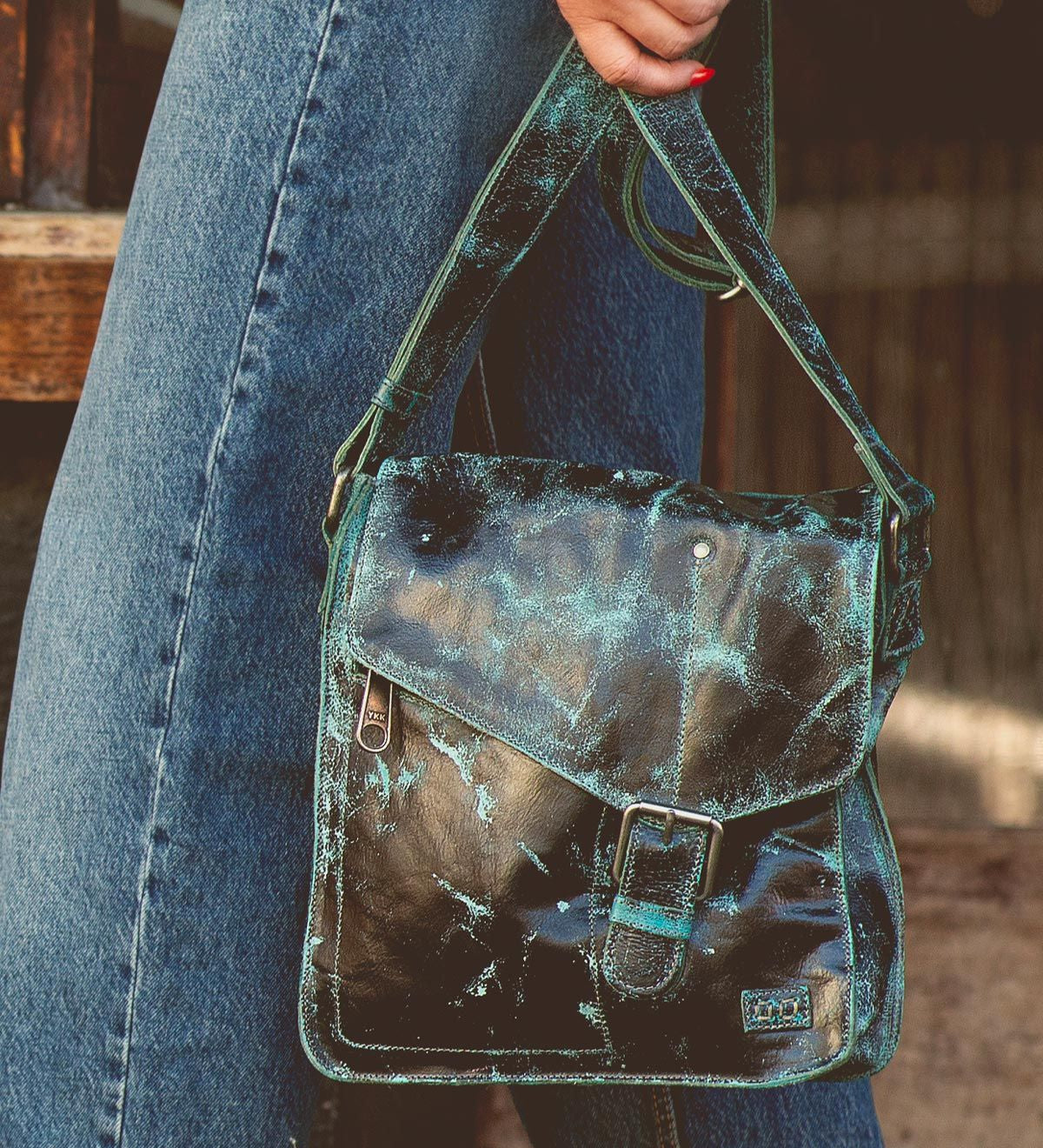 A person wearing blue jeans holds a Bed Stu Venice Beach handmade leather crossbody bag with an aged, distressed green and black leather finish and a buckle on the front flap.