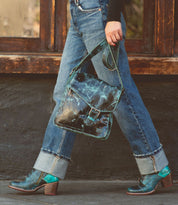A person walking outdoors, holding a Bed Stu Venice Beach handmade leather crossbody bag and wearing cuffed blue jeans along with matching green and brown heeled shoes.