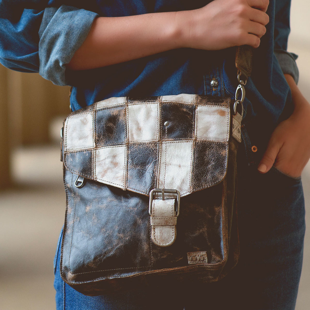 A person wearing a blue denim outfit is holding a Venice Beach II leather shoulder bag by Bed Stu with a checkered front panel and buckle closure.