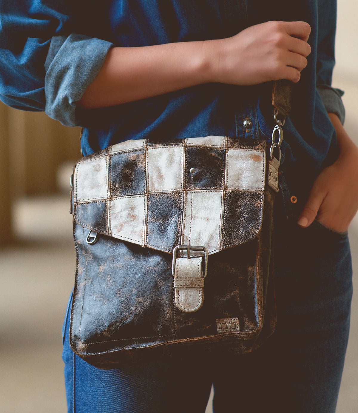 A person wearing a blue denim outfit is holding a Venice Beach II leather shoulder bag by Bed Stu with a checkered front panel and buckle closure.