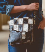 A person wearing a blue denim outfit is holding a Venice Beach II leather shoulder bag by Bed Stu with a checkered front panel and buckle closure.