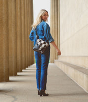A person with long blonde hair walks away from the camera, wearing a blue denim outfit and carrying the Venice Beach II by Bed Stu, a functional bag with a checkered front panel. They are in an outdoor corridor with tall columns, reminiscent of Venice Beach II.