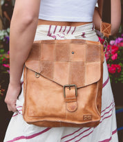 Person wearing a striped skirt and white crop top, carrying a functional bag with a Bed Stu Venice Beach II featuring a checkered front panel and buckle closure on their shoulder. Flowers are visible in the background.