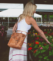 A woman in a white tank top and a striped skirt, carrying the Venice Beach II bag by Bed Stu with a checkered front panel, is admiring vibrant flowers.