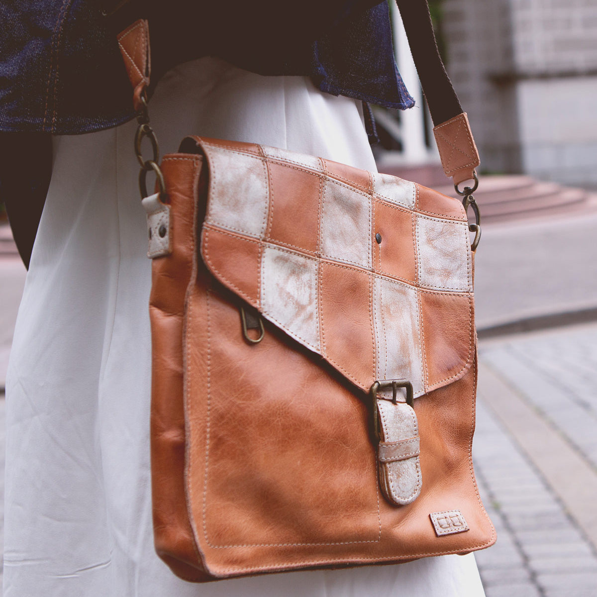 A person is carrying a Bed Stu Venice Beach II with a buckle closure and a functional checkered front panel. The background includes a paved walkway and part of a building, evoking the laid-back vibe of Venice Beach.