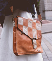 A person is carrying a Bed Stu Venice Beach II with a buckle closure and a functional checkered front panel. The background includes a paved walkway and part of a building, evoking the laid-back vibe of Venice Beach.