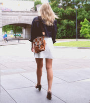 A woman with blond hair, wearing a white dress and denim jacket, walks away on a paved path with her Bed Stu Venice Beach II, featuring a checkered front panel. A bridge and greenery are in the background.