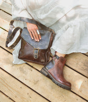 A person seated on wooden planks is wearing a white skirt, brown leather boots, and an adjustable finger brace while holding the Venice Beach crossbody bag from Bed Stu, renowned for its quality craftsmanship.