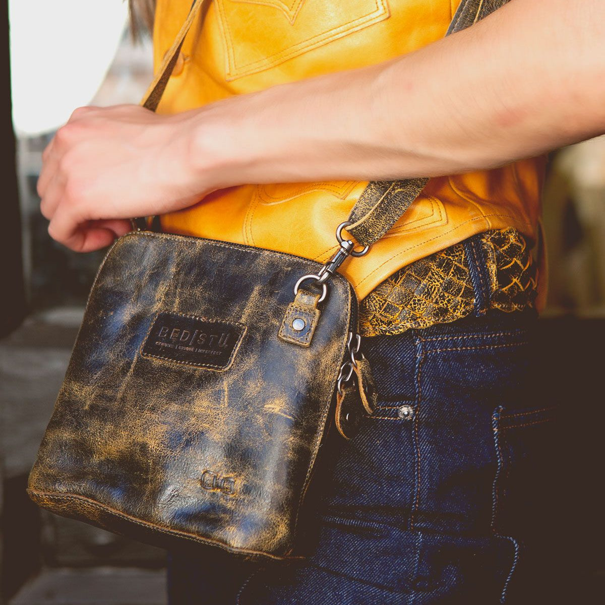 Close-up of a person wearing the Bed Stu Ventura leather crossbody bag and a yellow top. The bag features zippered pockets with the brand label and clasp visible. Only the person's torso and arm are shown.