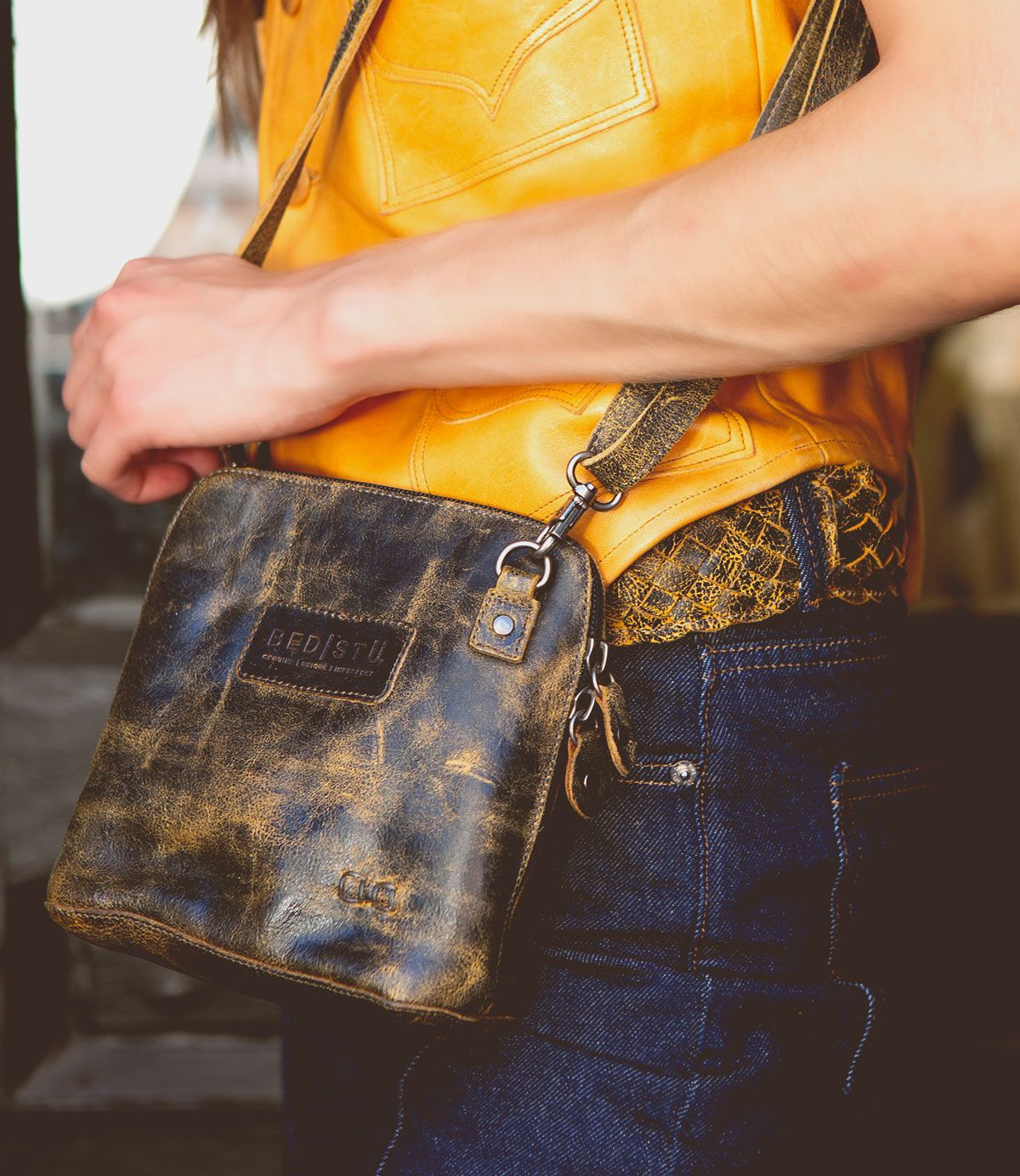Close-up of a person wearing the Bed Stu Ventura leather crossbody bag and a yellow top. The bag features zippered pockets with the brand label and clasp visible. Only the person's torso and arm are shown.