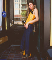 A woman wearing a yellow top and blue jeans leans against a black door with an ADT sticker. She carries a Bed Stu Ventura leather gem crossbody bag with zippered pockets and wears brown boots. Buildings and trees are visible in the background.