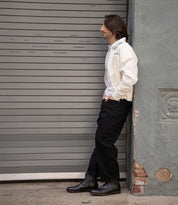 A man with shoulder-length hair leans against a gray wall in front of a closed metal shutter, wearing a white jacket, black pants, and Bed Stu Verrocchio handcrafted leather men’s boots.