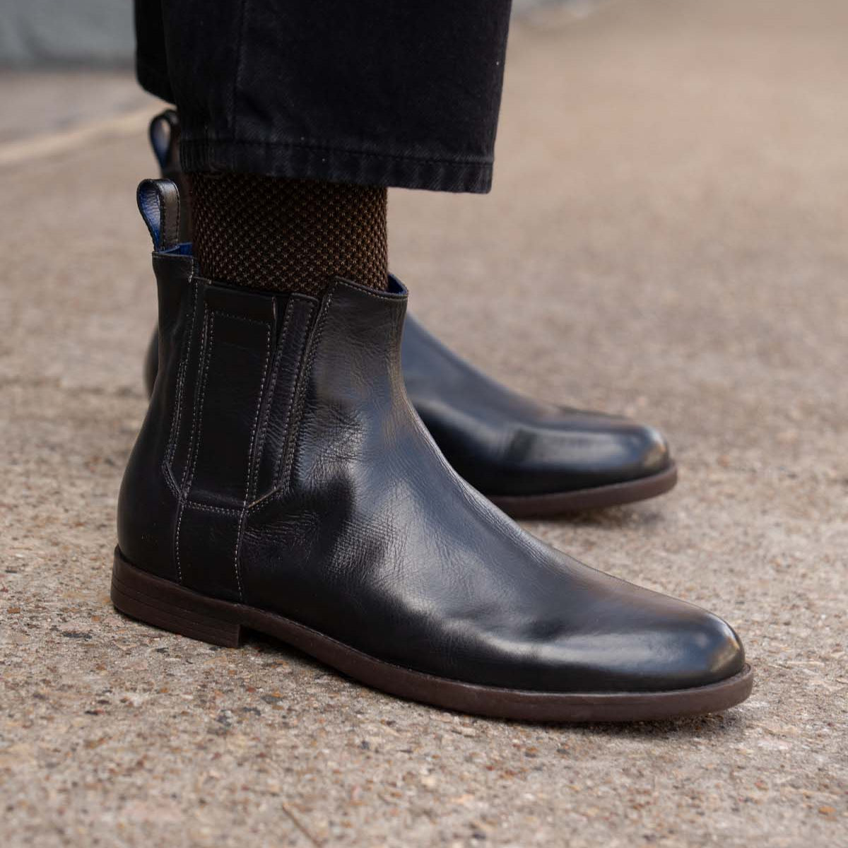 A person wearing Bed Stu Verrocchio black handcrafted leather slip-on boots and black pants stands on a concrete surface.