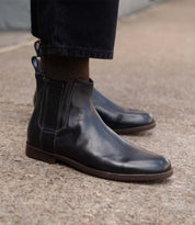 A person wearing Bed Stu Verrocchio black handcrafted leather slip-on boots and black pants stands on a concrete surface.